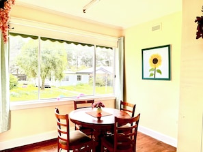 Un comedor con una mesa redonda y sillas, una ventana con vista a una casa y follaje, y un cuadro de un girasol en la pared.