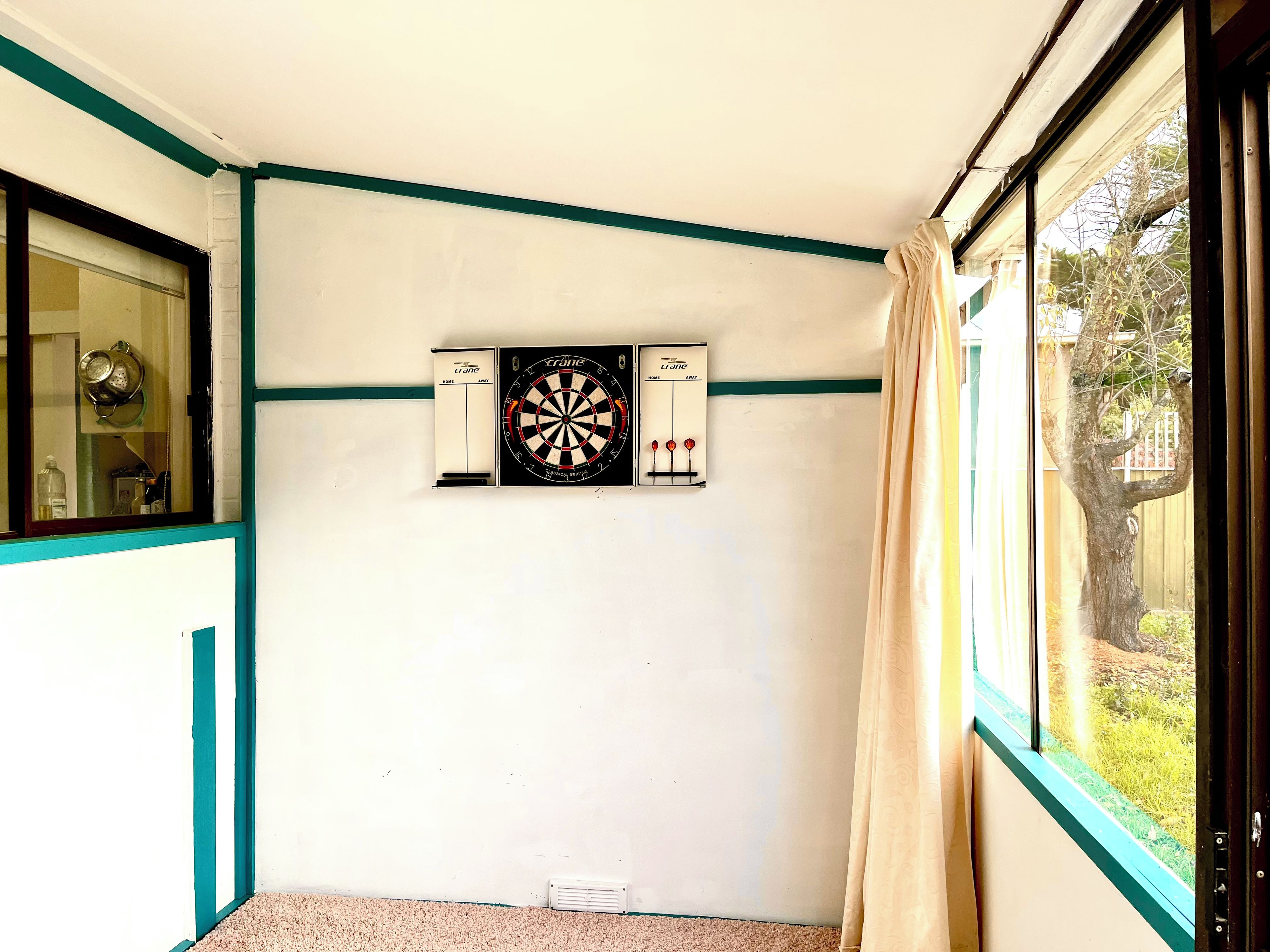 Een kamer met een dartbord, een raam met gordijn en een deur met blauwgroene afwerking.
