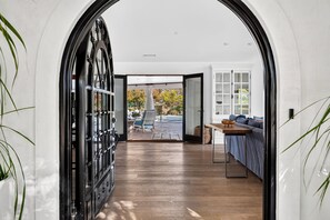 Un pasillo que conduce a una habitación con piso de madera, pared blanca y vista a un área exterior con piscina y sillas.