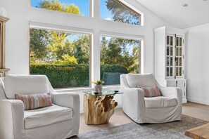 Una sala de estar moderna con dos sillones blancos, una mesa de centro de cristal y vistas a un jardín a través de amplios ventanales.