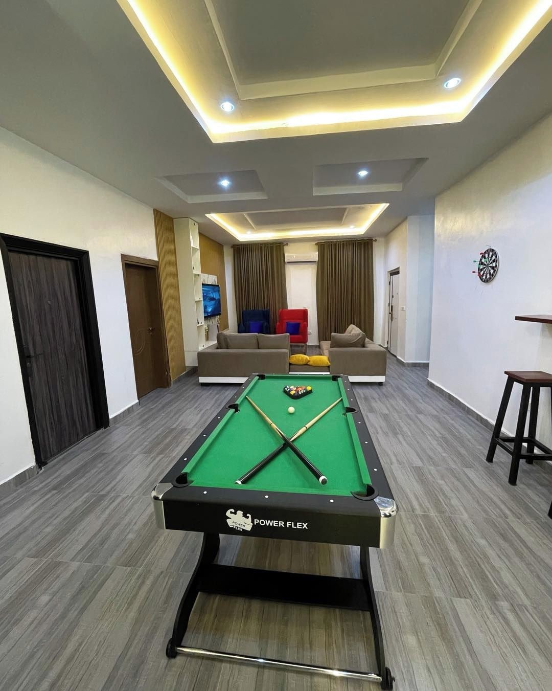 Une table de billard portant la mention « POWER FLEX », un téléviseur et un jeu de fléchettes dans une pièce au design moderne, comprenant un éclairage encastré et un canapé.