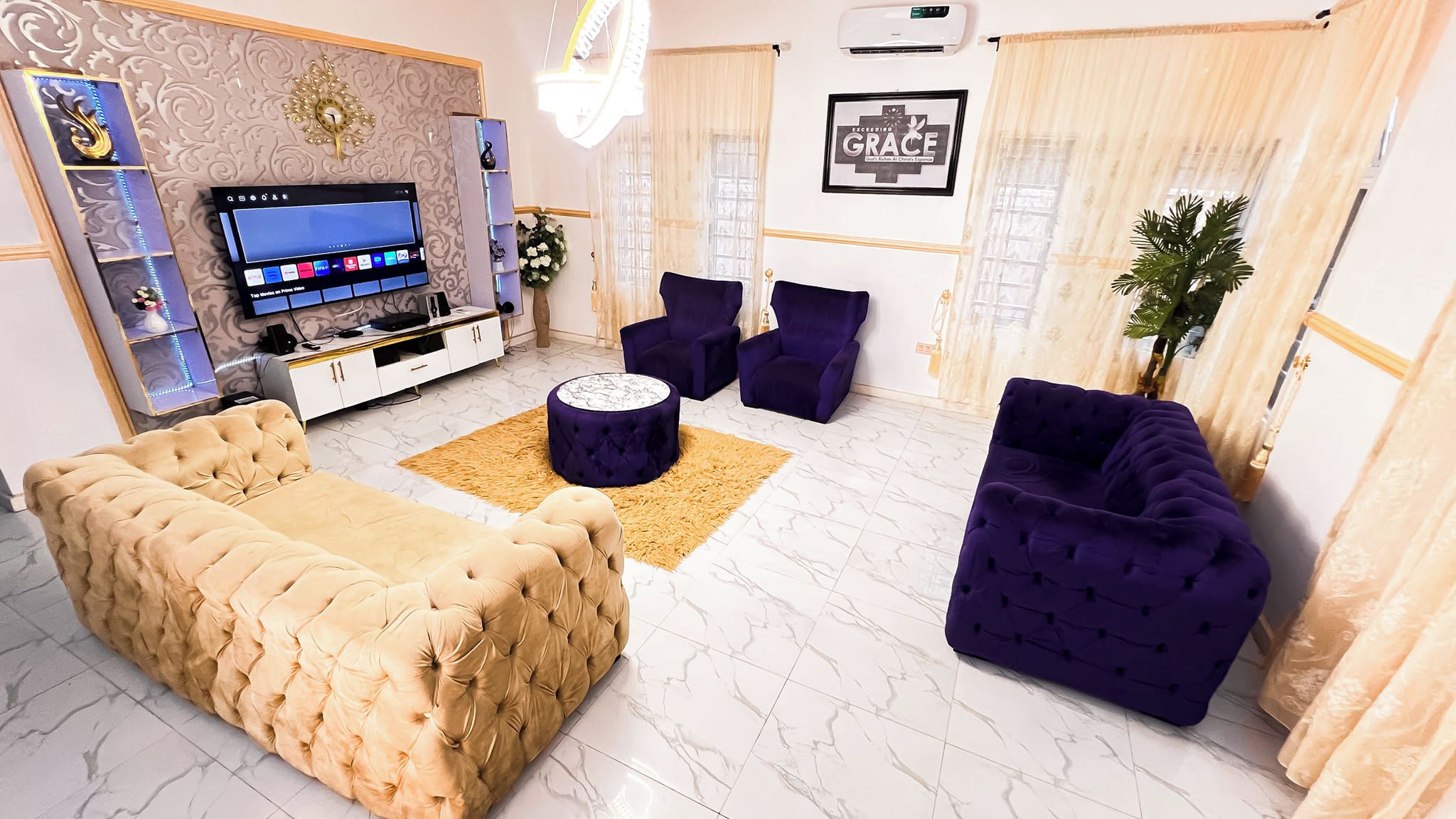 Ein Wohnzimmer mit einem beigefarbenen Sofa, zwei violetten Sesseln, einem Couchtisch und einem Fernseher auf einem weißen Schrank.