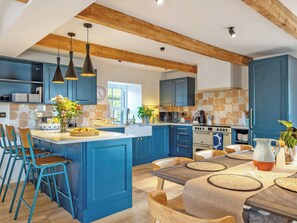 A modern kitchen with blue cabinetry, a central island, bar stools, and a dining table with placemats. - Fairlie Ward Cottage (Fairlie)