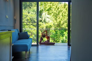 A room with a blue sofa, a person doing yoga, and a view of greenery through a large window. - The Source (Ko Pha-ngan)