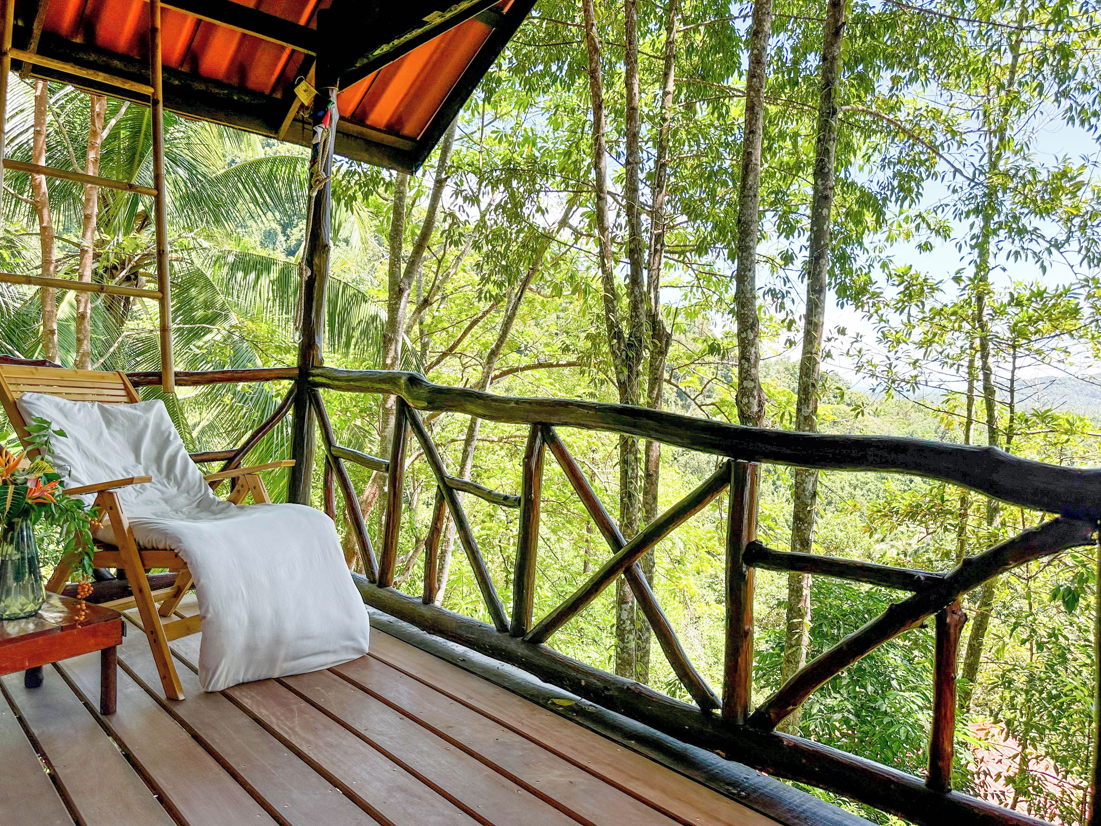 Terraza de madera con una única silla de descanso y una mesa, con vista a un bosque frondoso.
