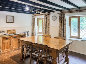 A dining area with a wooden table and chairs, a wooden cabinet, a clock on the wall, and windows with curtains. - 4 bedroom accommodation in Bickington (Bickington)