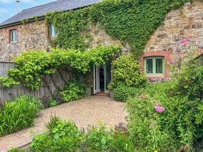 A stone building with a brick section, a wooden fence, and a paved garden area with various plants and flowers. - Ostler's Cottage - UK52192 (Beaworthy)