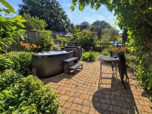 Eine Terrasse mit Whirlpool, Tisch und Stühlen, umgeben von üppiger Bepflanzung und einem Holenzaun.