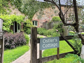 A sign for Ostler's Cottage with a brick wall and greenery in the background. - Ostler's Cottage - UK52192 (Beaworthy)