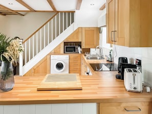 A modern kitchen with wooden cabinets, a built-in washing machine, a microwave, and a toaster. - Ostler's Cottage - UK52192 (Beaworthy)