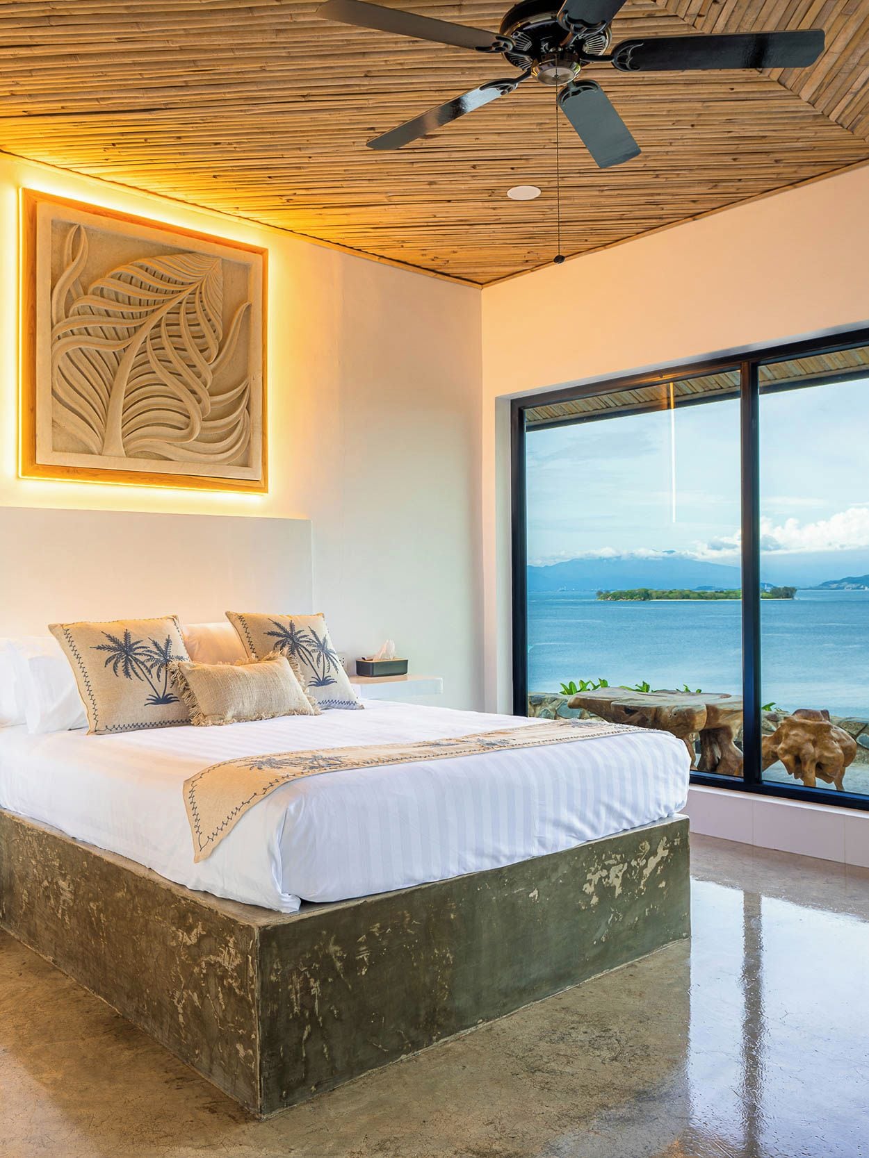 Un dormitorio con una cama grande, un ventilador de techo, techo de madera, una obra de arte enmarcada y vista al mar a través de un ventanal amplio.