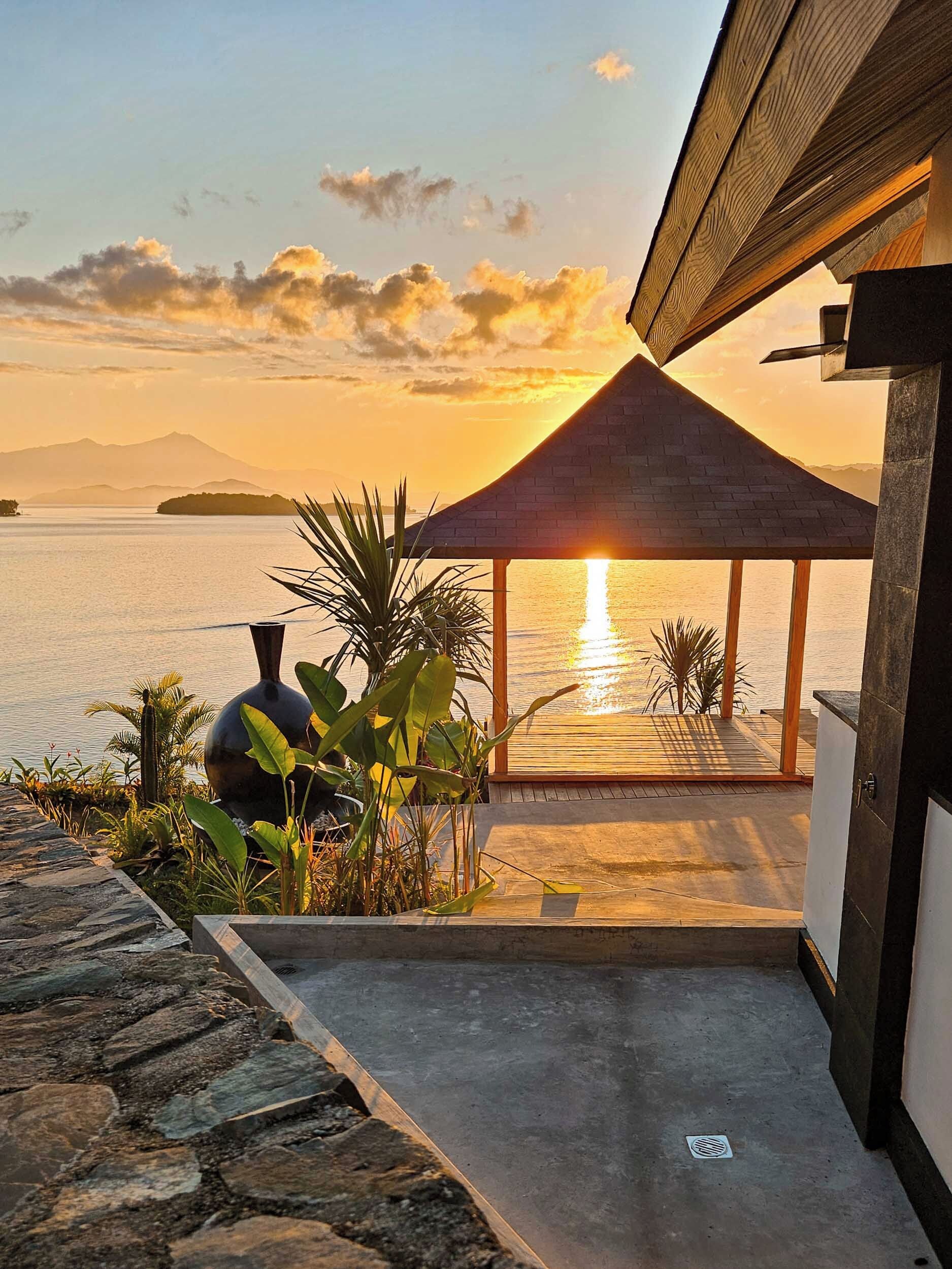 Un espacio al aire libre techado con vistas a la puesta de sol sobre el agua, un sendero de piedra y plantas tropicales.
