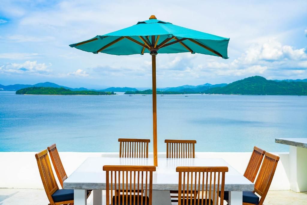Una mesa con cuatro sillas y un paraguas, con vista a un mar tranquilo y islas lejanas.