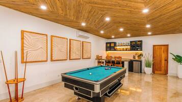 A pool table with a billiard cue and balls, a bar area with bottles, and a wooden ceiling.