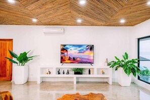 Un saló modern amb sostre de fusta, un televisor pla gran muntat a la paret, prestatges blancs amb objectes decoratius i plantes grans en testos.