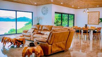 A spacious living area with a leather sofa, dining table, chairs, and a large window offering a view of a lake and mountains.