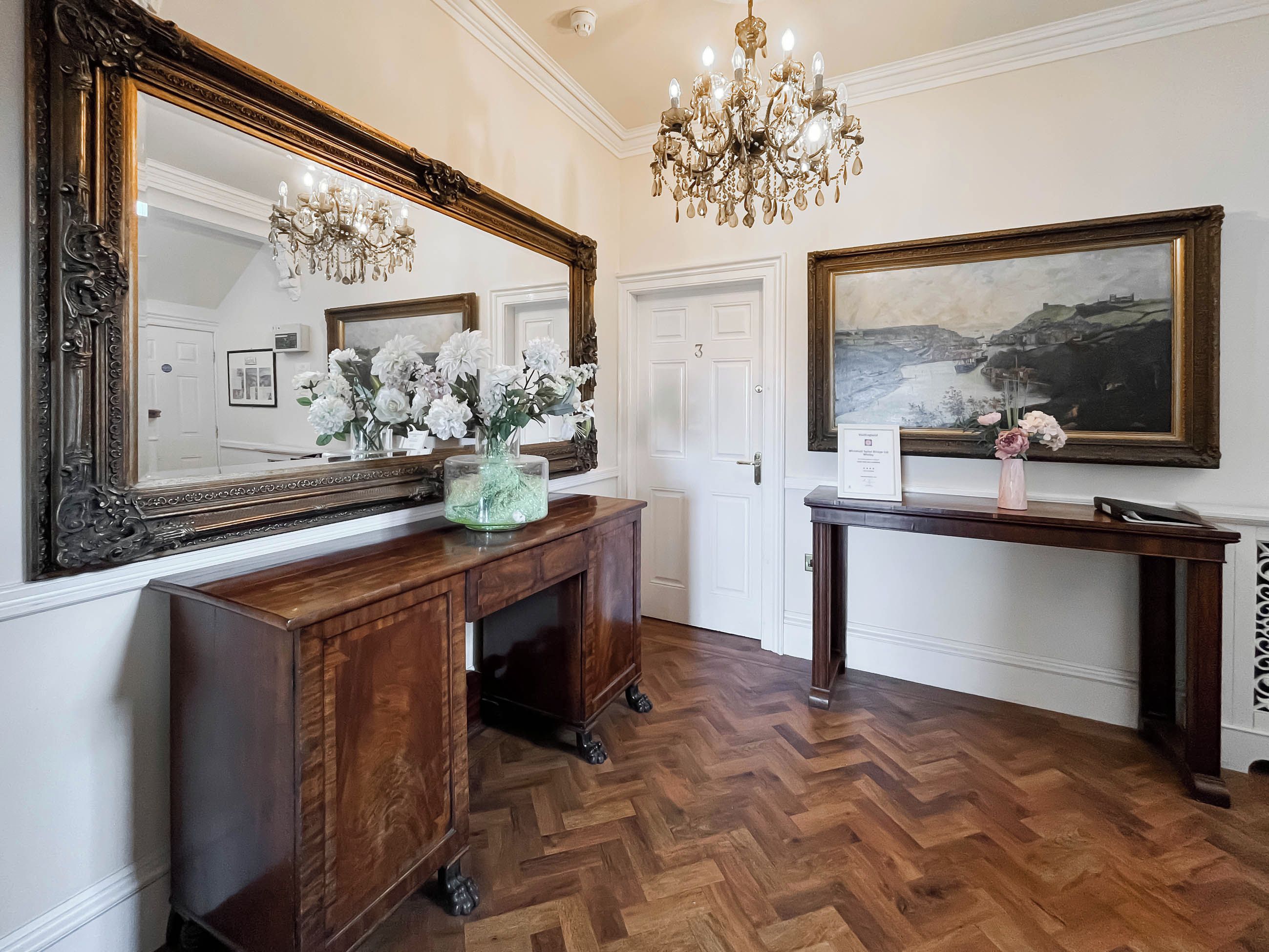 A room with a large ornate mirror, a wooden console table with a vase of flowers, a painting on the wall, and a herringbone-patterned floor.