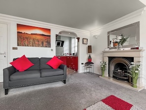 A living room with a grey sofa, red cushions, a fireplace, and a painting of a field. - The Genoa Suite at Whitehall (Whitby)