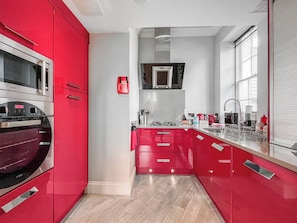 A modern kitchen with red cabinetry, stainless steel appliances, and a central island with a sink and countertop. - The Genoa Suite at Whitehall (Whitby)