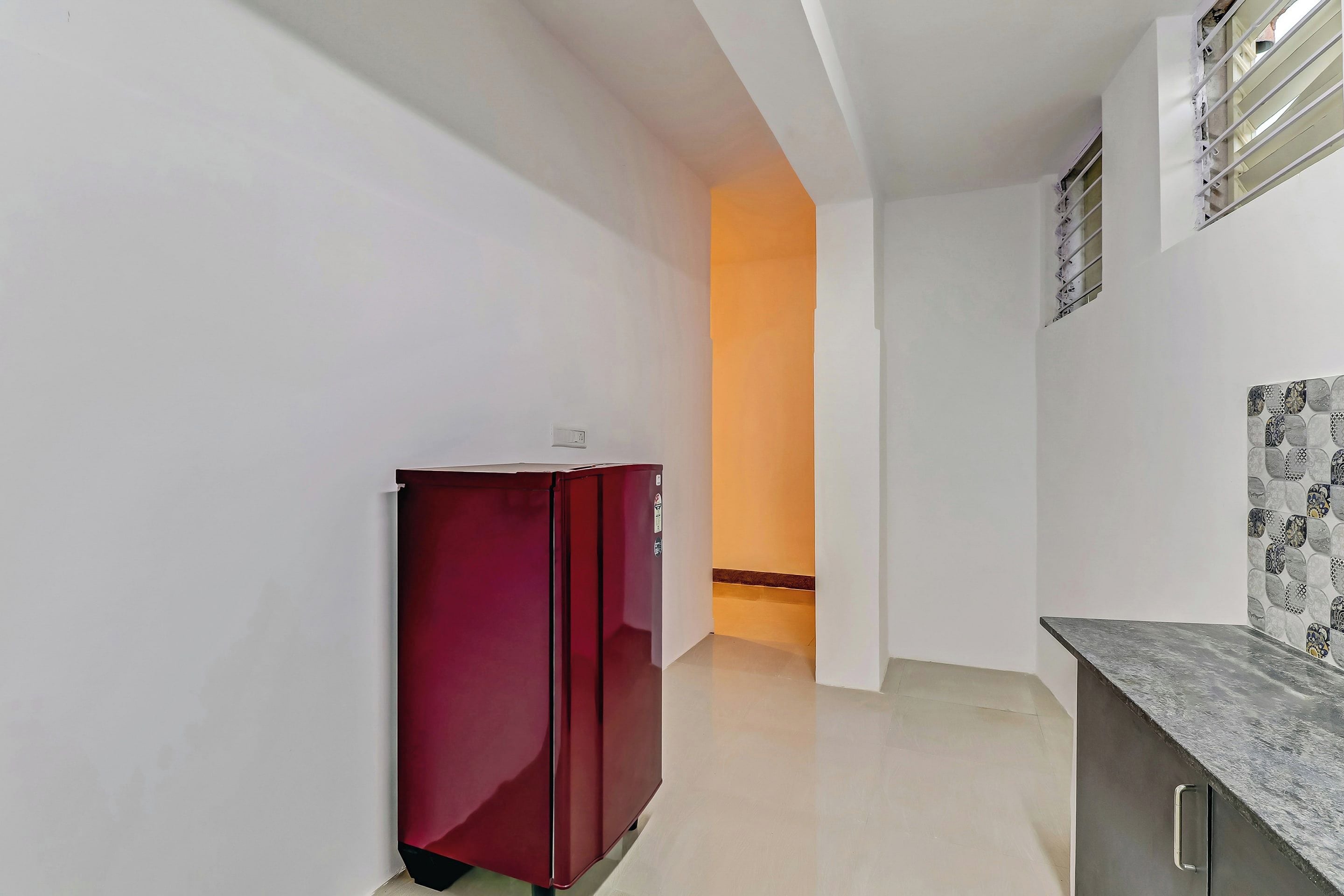 A narrow hallway with a red refrigerator, a white wall with a decorative tile pattern, and a door leading to another room.
