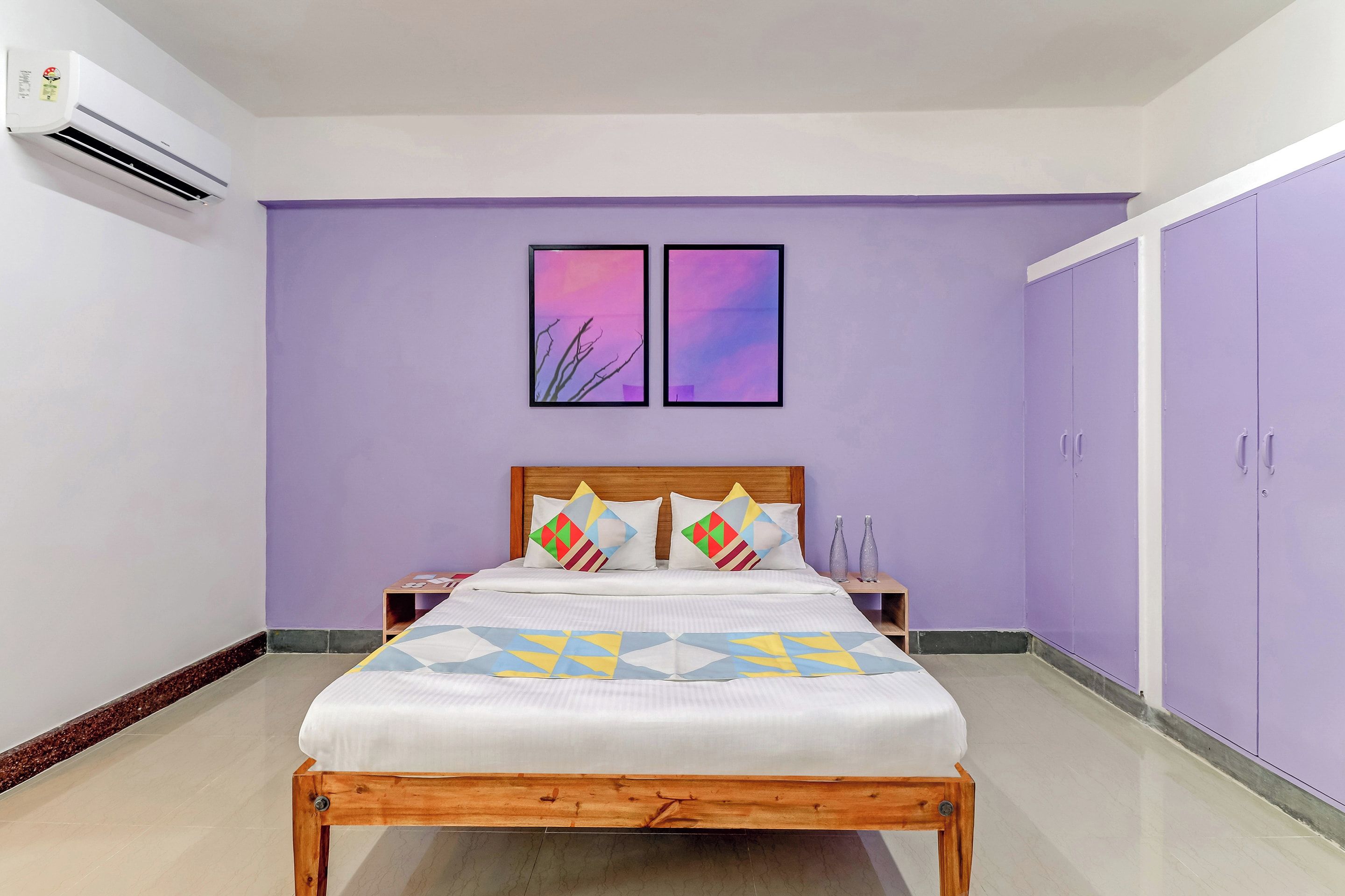 Un dormitorio con cama, dos mesitas de noche, cabecera de madera, pared morada, dos cuadros enmarcados y un armario.