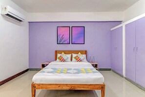 Un dormitorio con cama, dos mesitas de noche, cabecera de madera, pared morada, dos cuadros enmarcados y un armario.