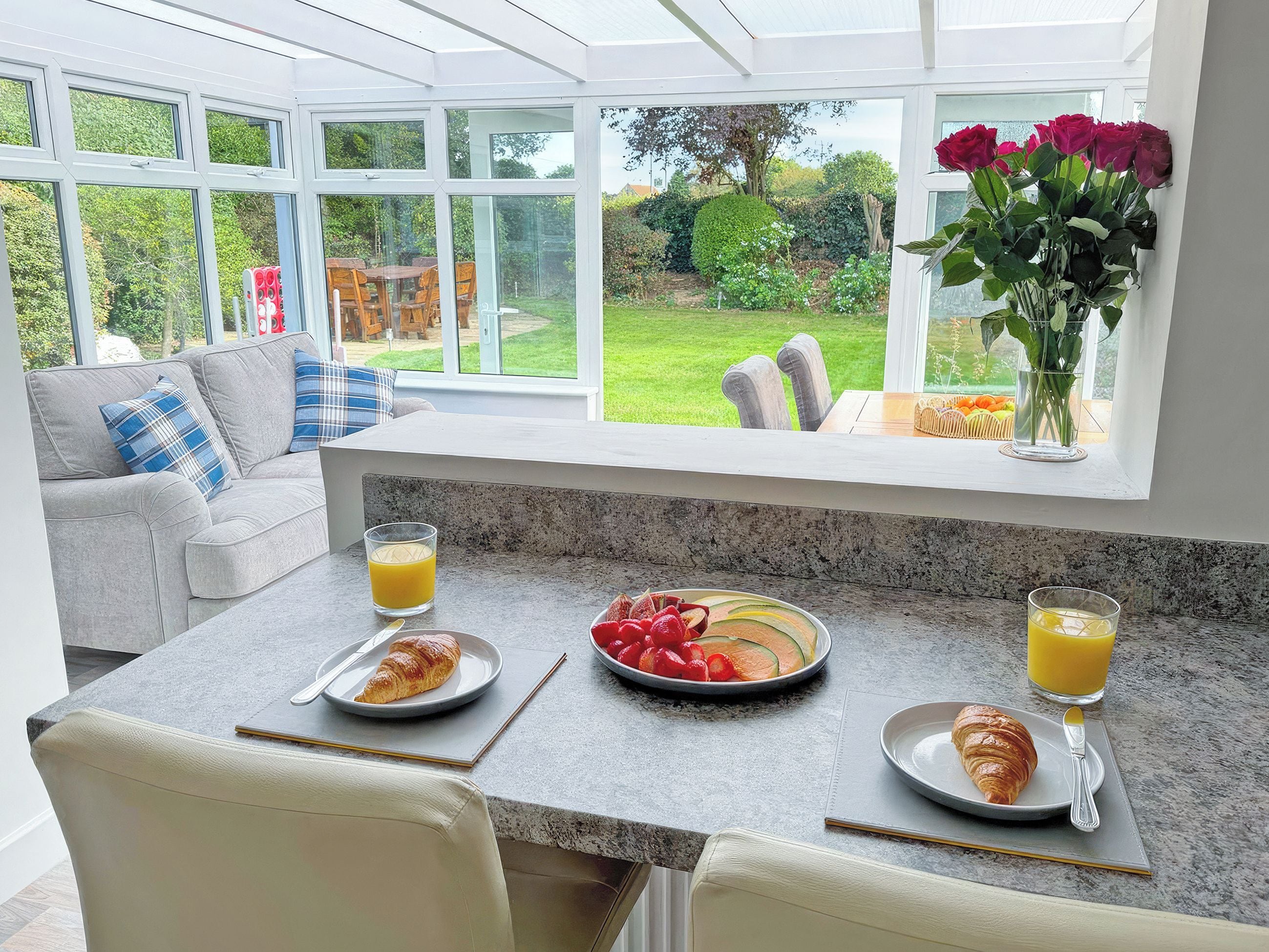 Un plateau de petit-déjeuner avec des croissants, des fruits et du jus d’orange, disposé sur un comptoir de cuisine, avec vue sur un jardin.