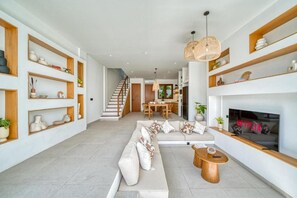 A modern living room with a sofa, coffee table, and fireplace. There are wooden shelves and a staircase in the background. - Kembali 2BR Private Pool Villa Balangan (Badung)