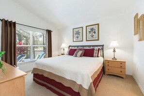 Ein Schlafzimmer mit einem Bett, zwei Nachttischlampen, einem Nachttisch, einem Fenster mit Vorhängen und zwei gerahmten Bildern an der Wand.