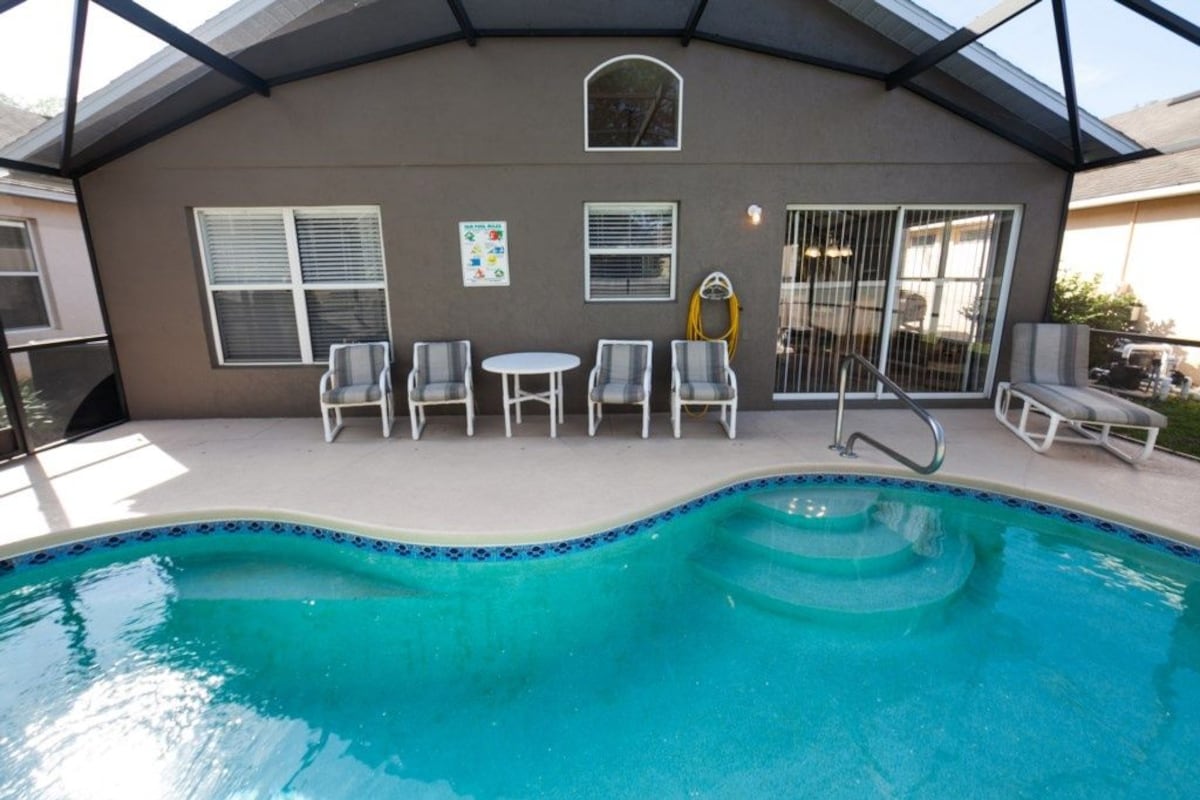 Ein Schwimmbecken mit einer gefliesten Kante, umgeben von weißen Liegestühlen und einem kleinen Tisch.