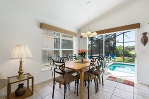 Ein Essbereich mit Holztisch und -stühlen, einer Lampe, einer Blumenvase und Blick auf einen Swimmingpool durch große Fenster.