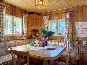 A wooden cabin interior with a round dining table, benches, and a view of the mountains through the windows. - Schneehitzalmhütte by Interhome (Wörschach)