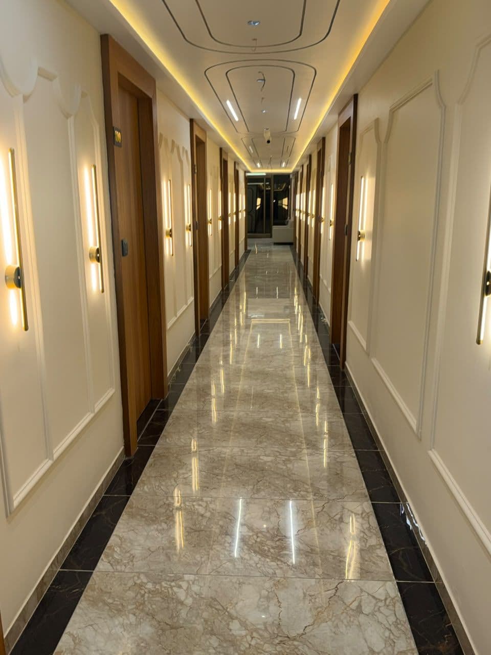 Un pasillo de hotel con puertas a ambos lados, suelo de mármol y apliques de pared que proporcionan luz.