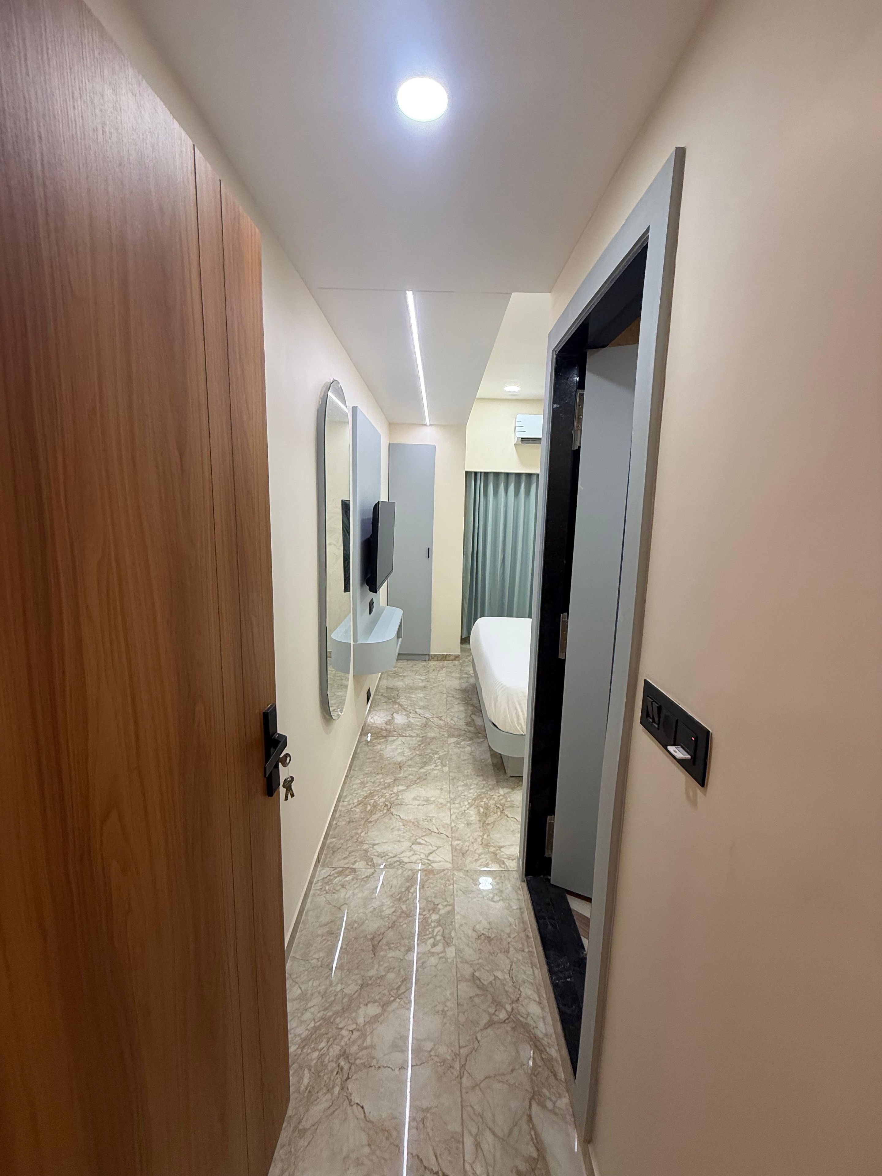 Pasillo de hotel con puerta de madera, baño con espejo y lavabo, cama y lámpara en el techo.