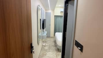 Un couloir d’hôtel avec une porte en bois, une salle de bain avec un miroir et un lavabo, un lit et un luminaire au plafond.