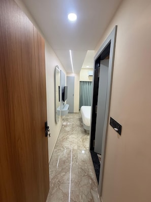 Passadís d'hotel amb una porta de fusta, un bany amb mirall i rentapès, un llit i una làmpada al sostre.