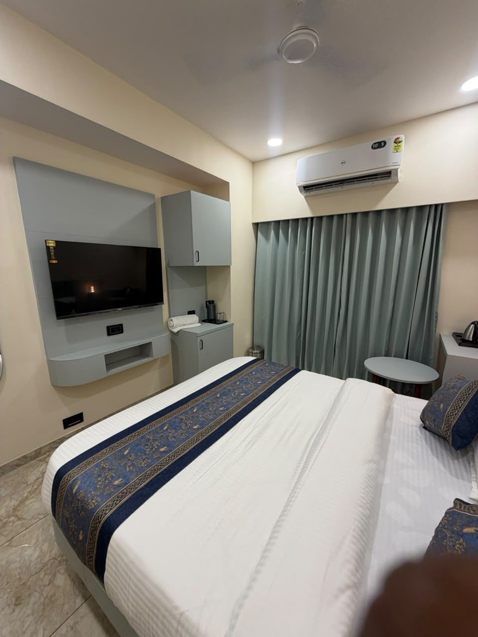 A hotel room with a bed, a television, a desk, a chair, an air conditioning unit, and a window with curtains.