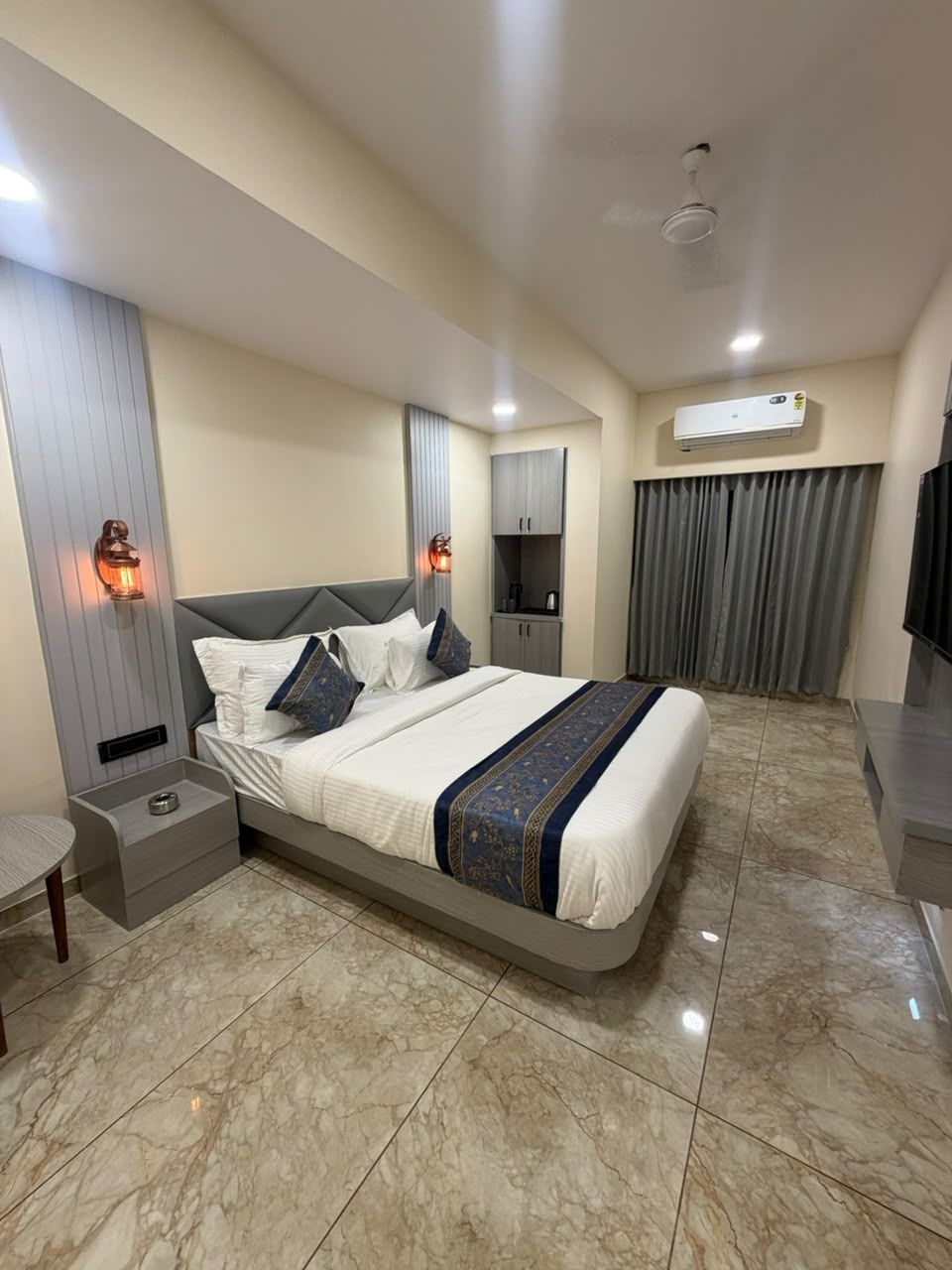 A hotel room with a large bed, a bedside table, a nightstand, a wall-mounted air conditioner, and a window with curtains.