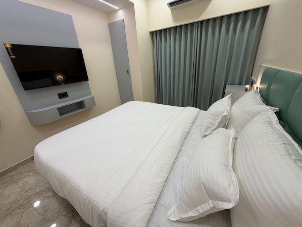 Habitació d'hotel amb un llit, una televisió de pantalla plana penjada a la paret, un panell de capçalera gris i una finestra amb cortines.