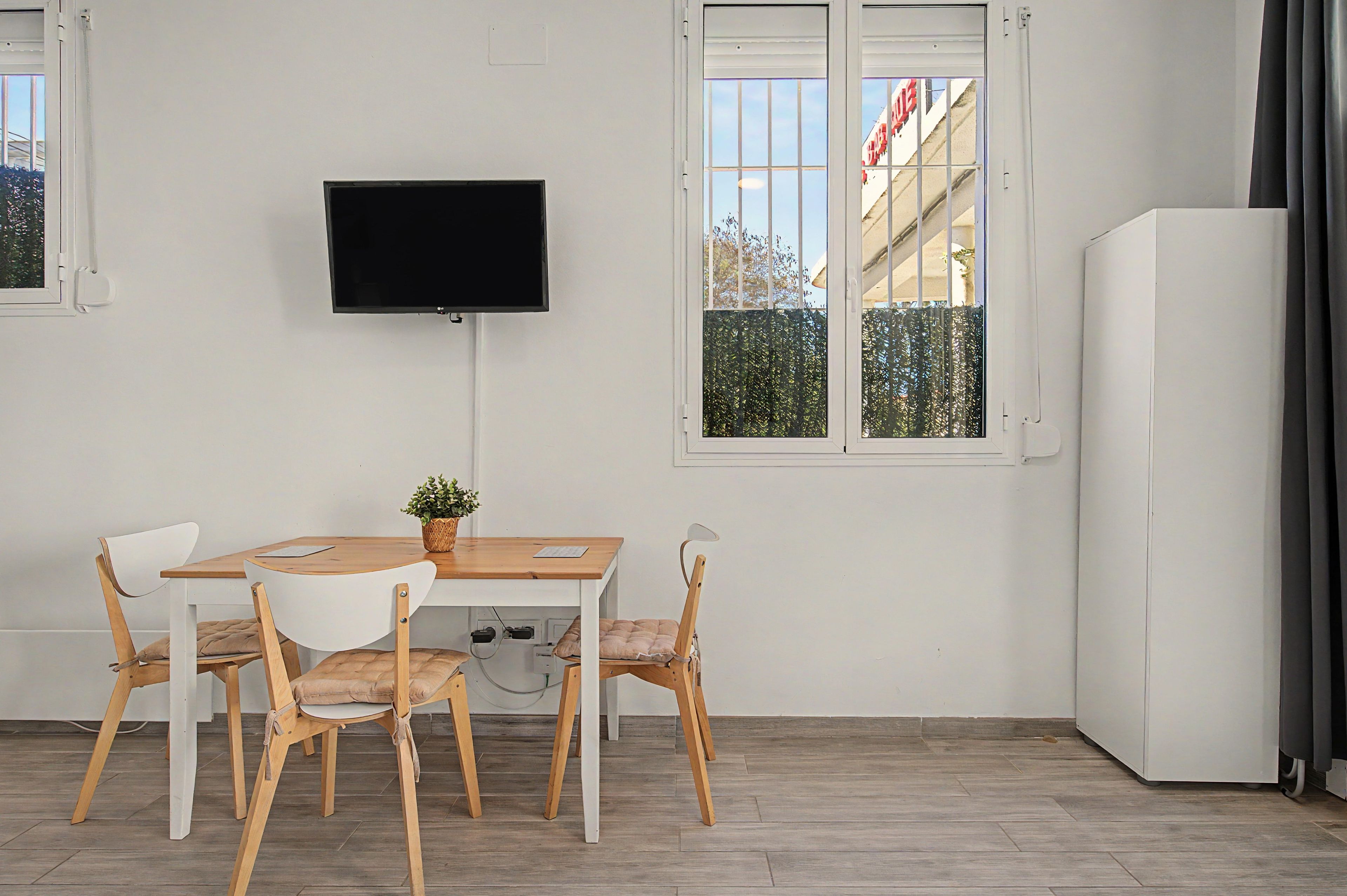 Una habitación con una mesa de madera, dos sillas, un televisor montado en la pared y un ventanal con vistas a edificios y árboles.