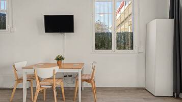 Una habitación con una mesa de madera, dos sillas, un televisor montado en la pared y un ventanal con vistas a edificios y árboles.