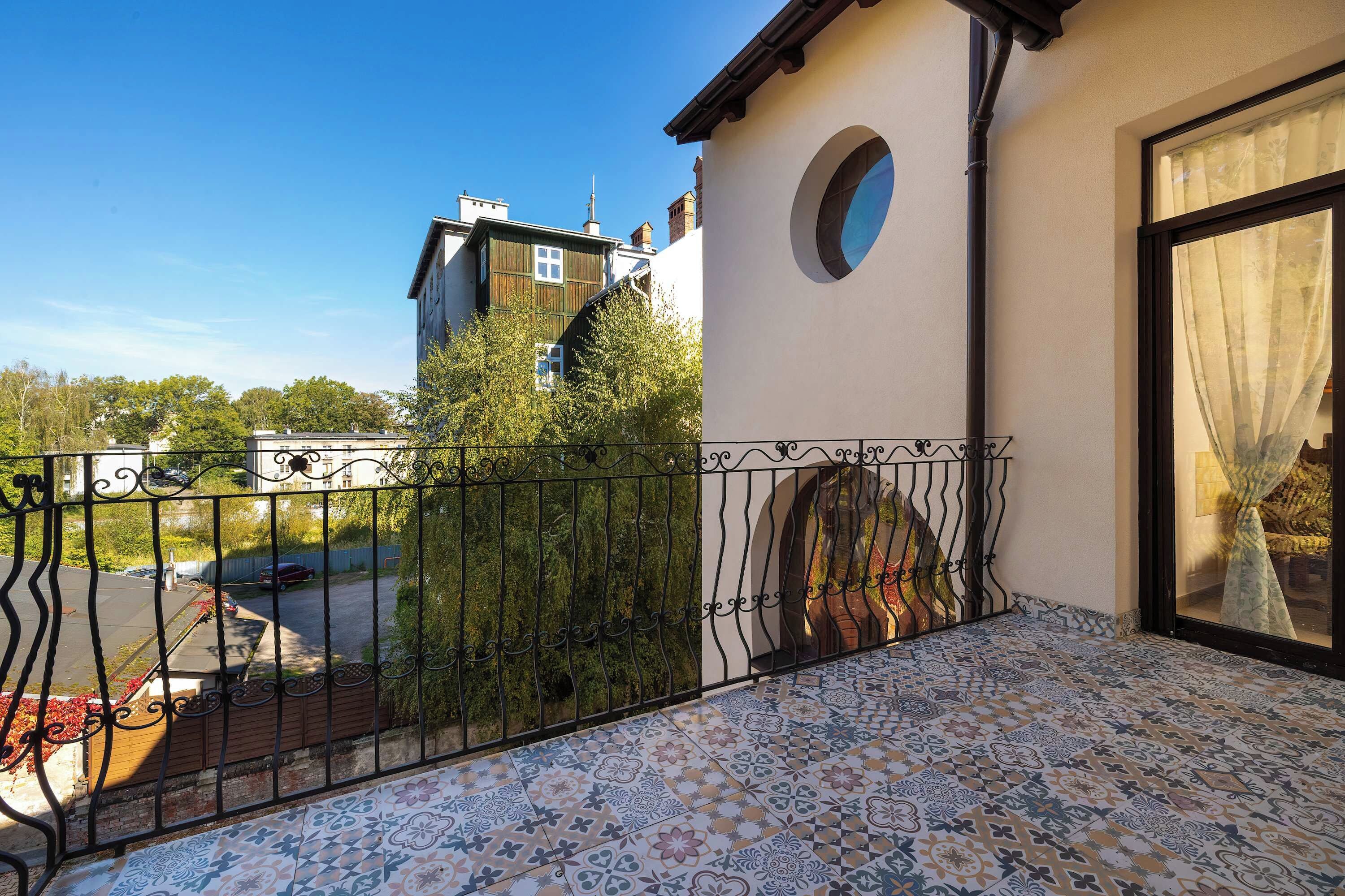 A balcony with patterned floor tiles, a wrought iron fence, and a view of a street with parked cars and buildings.