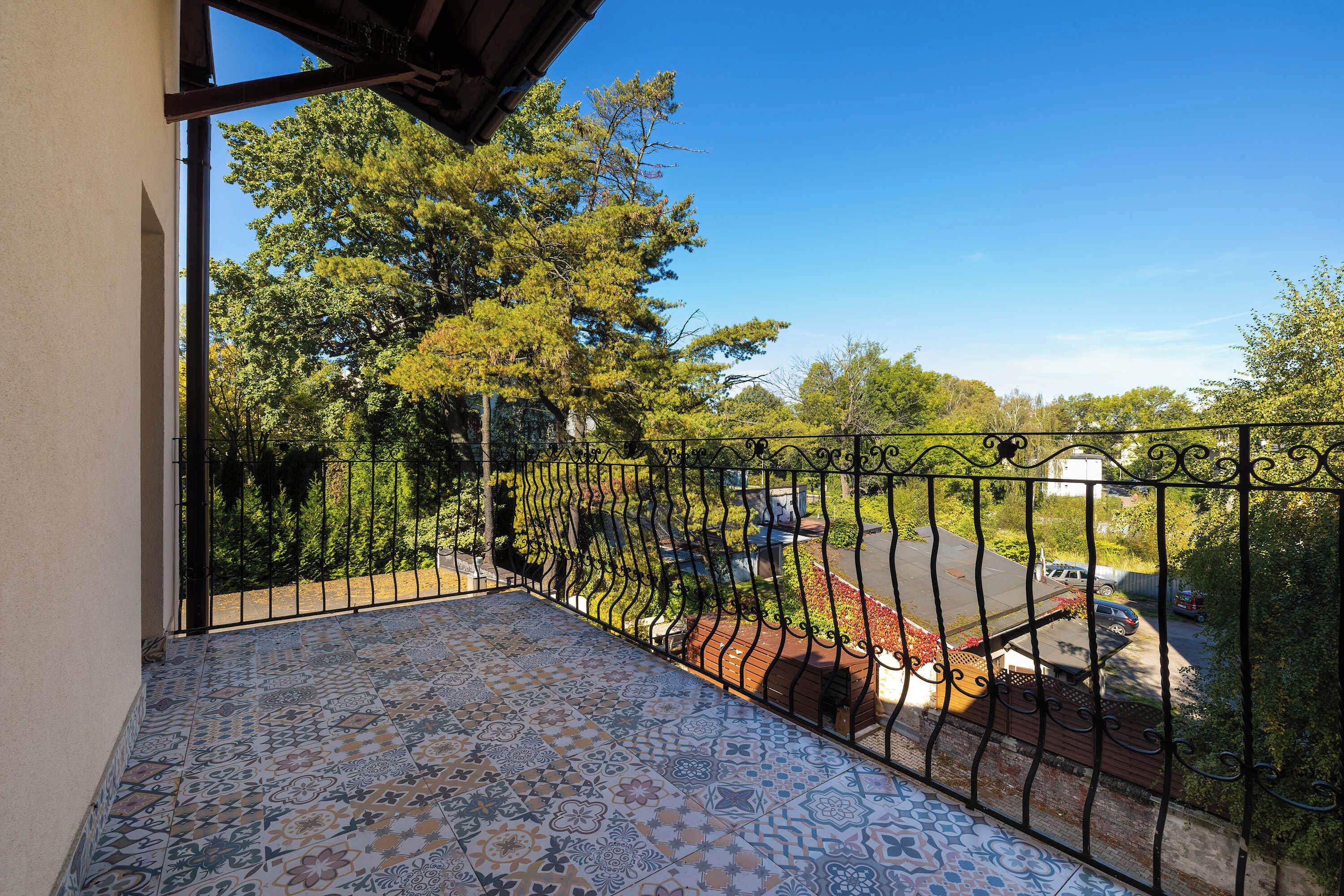 A balcony with patterned floor tiles, a metal railing, and a view of trees and parked cars.