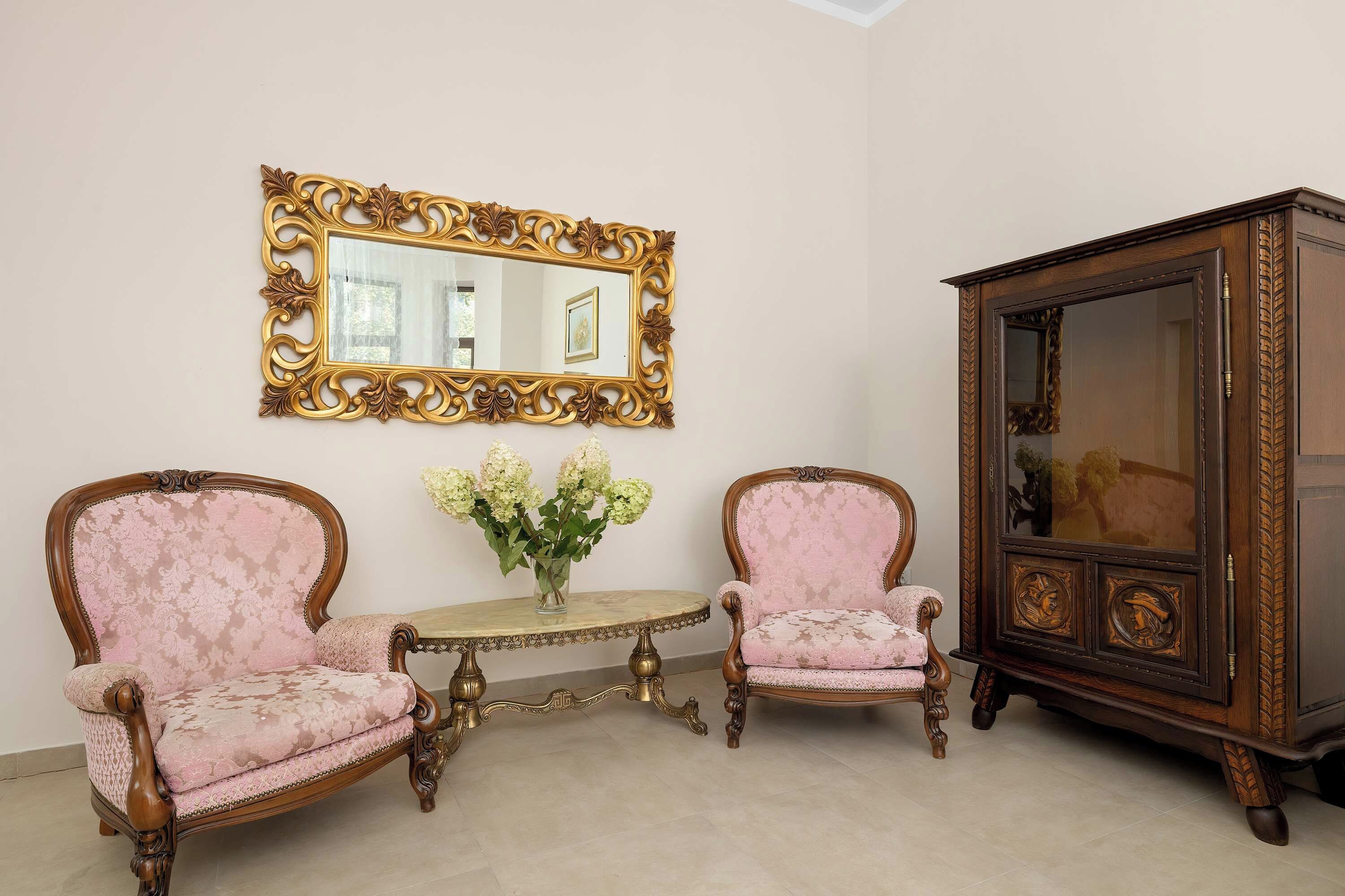Una stanza con due poltrone decorate, un tavolino, uno specchio con una cornice elaborata e un mobile con anta in vetro.