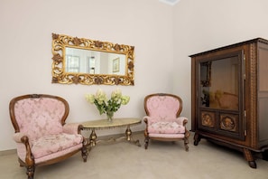 Um quarto com duas cadeiras ornamentadas, uma mesinha, um espelho com moldura elaborada e um armário com porta de vidro.