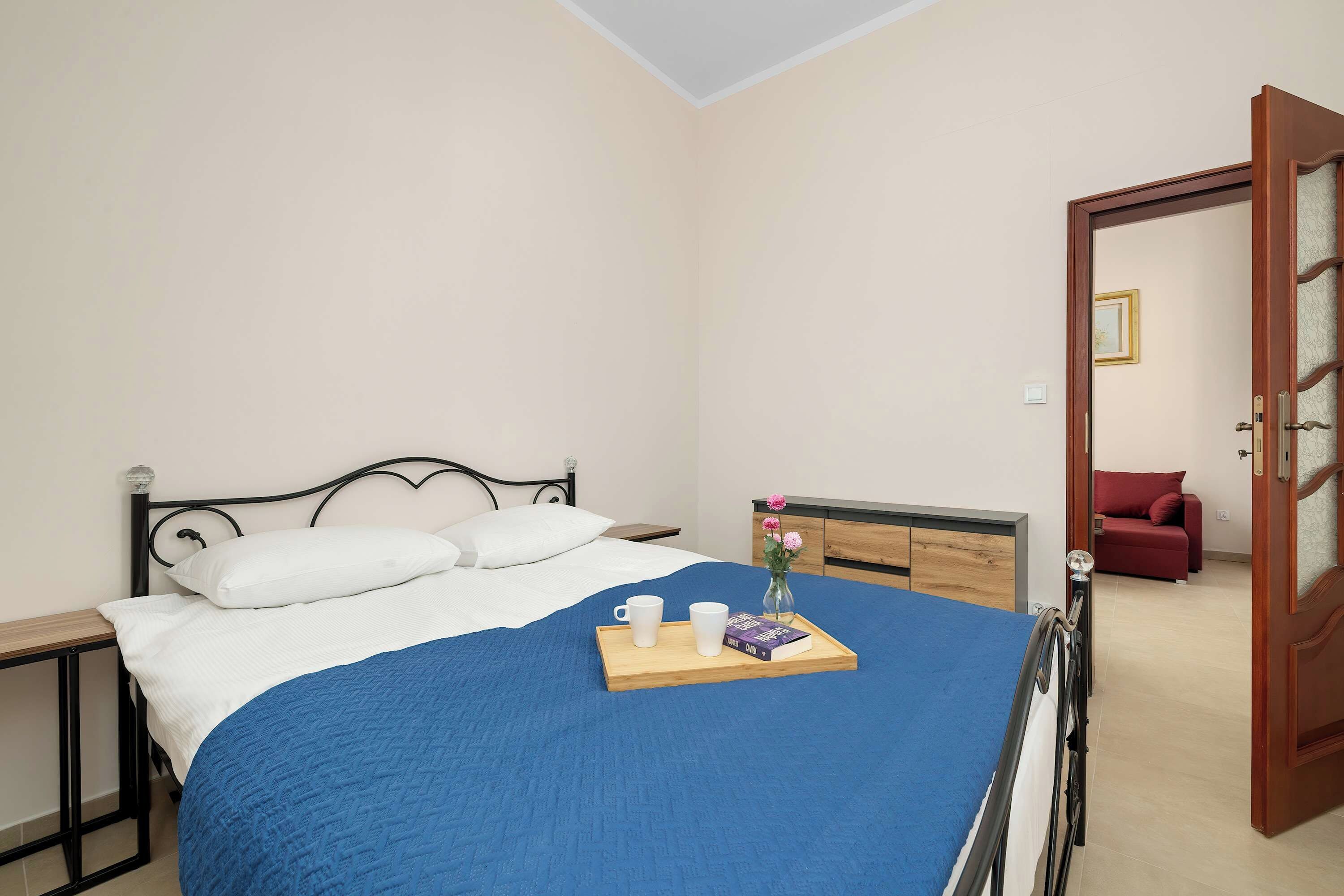 A neatly made bed with a blue blanket, a bedside table with a tray of books and a cup, and a red sofa in the adjacent room.
