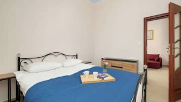 A neatly made bed with a blue blanket, a bedside table with a tray of books and a cup, and a red sofa in the adjacent room.