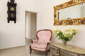 Una habitación con una silla de estilo vintage, un reloj de pared, un espejo con detalles dorados ornamentales y un florero con flores sobre una mesa de mármol.