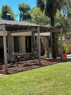 A covered outdoor seating area with tables and chairs, a hanging chair, and a pool in the background. - Charming B&B Retreat in Springs with Private Room and Shared Pool (Springs)