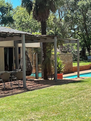 A covered outdoor seating area with a pool, surrounded by greenery and a brick wall.
